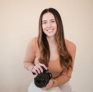 Woman photographer smiling at the camera wearing a long-sleeve tan shirt and holding a camera